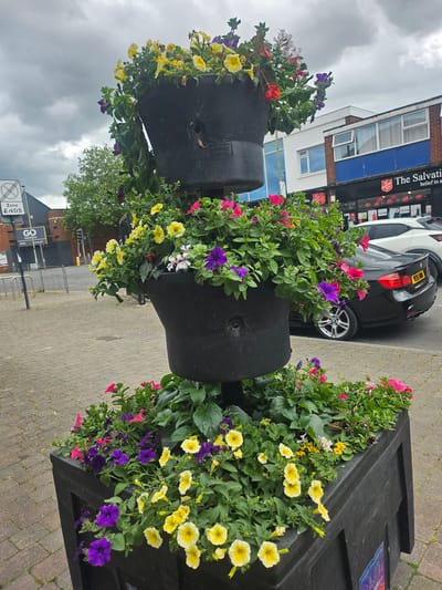 The image shows two large, dark-colored, urn-shaped planters filled with an assortment of colorful flowers including yellow, purple, and pink petunias and other varieties. The planters are situated on a brick-paved walkway. Behind the planters, a black and white car is parked near a building with the words "The Salvation" visible on the facade. A street with traffic lights and a street sign that reads "Zone Ends" can be seen in the background, along with trees and other buildings. The sky is overcast. The location is within Gloucester, United Kingdom.