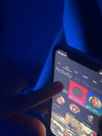 The image shows a person holding a smartphone displaying an application with an "Activity" tab selected. The screen displays content related to "All Favorite" activities, including entries such as "staker redeen," "new memory," and "collected daily." The screen also features profile pictures of individuals associated with these activities. The phone displays a time of "00:384." The surrounding environment appears to be dimly lit with a blue-ish hue. There is no indication of the city or country in the image.