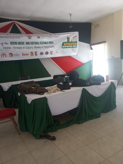 The image depicts an interior view of an event setup at Moi Girls High School in Eldoret, Kenya, within what would be categorized as Racecourse ward. The scene shows a large room with a light-colored tiled floor and a plain white ceiling featuring an exposed conduit and a single ceiling fan. A large window on the right side provides natural light.

In the background, a stage or backdrop is created using dark green, white, and red draped fabric, resembling a large tent or festive decoration, set against a dark fabric sheet. A prominent white banner is stretched across the top of this backdrop, advertising "THE KENYA MUSIC AND CULTURAL FESTIVALS 2025" with the theme "Threads of Culture, Weaves of Opportunity." The banner specifies the dates "30th Nov - 03rd Dec 2025" and the venue "Moi Girls High School - Eldoret," along with multiple sponsor logos including M-Pesa, Kenya Cultural Centre, TV47, and Pepsi.

Two rectangular tables are positioned in the foreground and midground, covered with white tablecloths and draped with additional green fabric that pools onto the floor. On the tables, several items are visible: a brown jacket or coat, a dark brown leather bag, and other light-colored fabric items on the left table. The right table holds multiple dark-colored bags, one of which appears to have white text (possibly "TRUTH"), a black rectangular object (possibly a laptop or audio device), and a red water bottle. Beneath the tables, on the tiled floor, a pair of dark shoes is visible. A single red-upholstered chair with wooden legs is partially visible in the lower-left foreground. No individuals are present in the scene, suggesting the setup is either awaiting attendees or has been recently vacated.