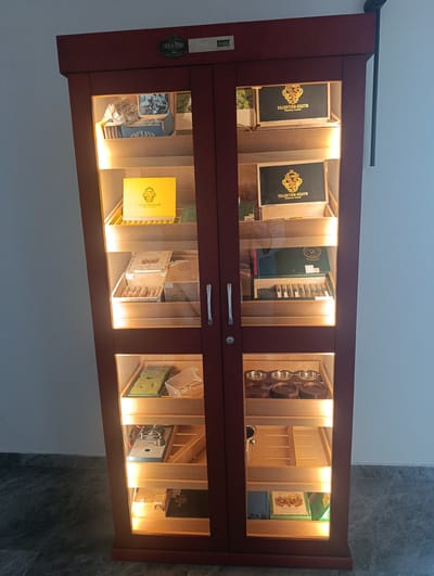 This is a media file showing a dark red humidor cabinet with glass doors. The cabinet is illuminated from within, highlighting various cigar boxes and accessories displayed on its shelves. The top shelf features a box labeled "ENCLAVE", and next to it, boxes of "VALENTINO SIESTO PREMIUM CIGARS". The second shelf from the top has another box of "VALENTINO SIESTO PREMIUM CIGARS" on the right and a bright yellow box with the "VALENTINO SIESTO" logo on the left. The third shelf displays several boxes of cigars, some in wooden boxes and others in cardboard packaging, including one labeled "ROBUSTO". The bottom shelves contain humidification elements, what appear to be cigar cutters or ashtrays, and some books. A digital display showing "50" and "38" is visible at the top of the cabinet, likely indicating temperature and humidity levels. The cabinet is situated in what appears to be an indoor setting, possibly a cigar lounge or a dedicated humidor room, given the context of the location as Čavori, Montenegro. The overall impression is one of organization and appreciation for premium cigars.