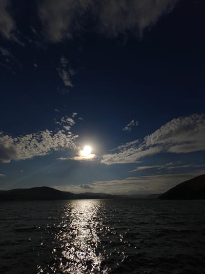 The image depicts a scenic view of a body of water reflecting the sun, with a silhouetted mountain range in the background. The sky is a deep blue, dotted with scattered white clouds. The sun is positioned to the left of the frame, its rays creating a shimmering path across the water. The mountains appear dark and are devoid of any visible detail. The water is calm, with small ripples. The location is specified as the city of New Bukhtarma, Kazakhstan. There are no people or any identifiable man-made structures in the image. The scene is one of natural landscape, characterized by water, sky, and mountains.