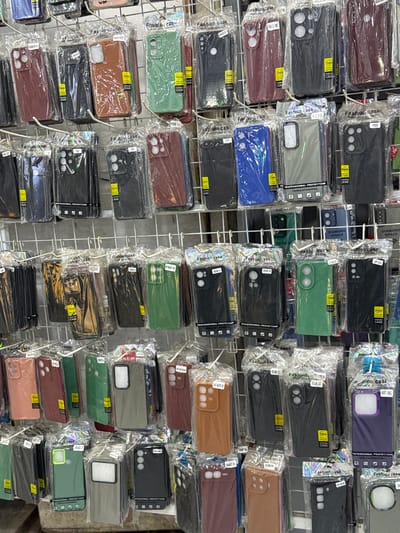 The image displays a retail display of mobile phone cases arranged on wire racks. The cases are packaged in clear plastic and feature various colors and designs, with some indicating specific phone models. The background consists of a plain wall. The image is cropped from a larger scene, and the provided crops offer closer views of different sections of the display. No people are visible in the image. The context suggests a shop or market stall selling phone accessories.
