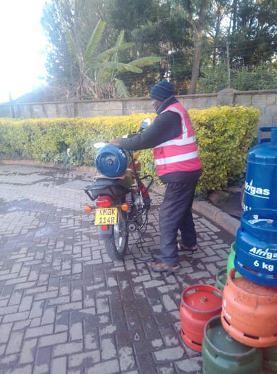 The image depicts an outdoor scene in Karai ward, Kenya, featuring a male individual, a motorcycle, and gas cylinders. A man, wearing a dark knitted beanie, a dark long-sleeved top, a red safety vest with two horizontal white reflective stripes, and dark trousers, stands next to a dark-colored motorcycle. He is positioned with his torso angled towards the front of the motorcycle, with both hands on a blue gas cylinder resting horizontally on the bike's rear rack, which has "32-12" inscribed on it. The motorcycle displays a yellow Kenyan license plate "KMGK 114R". To the right of the man, a stack of multiple gas cylinders is arranged, including blue cylinders clearly marked "Afrigaz 6 kg", alongside red, orange, and green cylinders. The ground is paved with wet, interlocking grey bricks. In the background, a light-colored concrete wall is topped with a dense green hedge. Beyond the hedge, various trees and large-leafed vegetation, including banana plants, are visible under a bright sky.