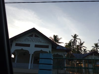 The image shows the exterior of a building in Rambongpayong, Indonesia. The building is a light color with a gabled roof and a decorated facade. The text on the building reads "MEUNASAH DESA TEUPIN RAYA KEC. PSG SIBLAH KRUENG". A fence runs in front of the building, and palm trees are visible in the background. The image appears to be taken from inside a vehicle. There are powerlines visible in the sky. The time of day appears to be dusk or dawn.