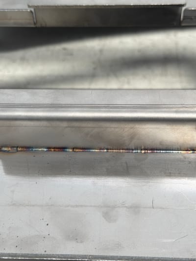 This is a close-up, high-angle shot of a metal surface with a visible weld seam. The subjects are metallic objects, possibly part of a structure or machine. The weld is the most prominent feature, running vertically down the center of the frame. It shows a progression of colors, from iridescent blues and purples to darker tones, indicating heat exposure during the welding process. The metal surface itself appears brushed or textured, with some light scratches and smudges visible. There's a distinct line or edge to the left of the weld, suggesting two separate pieces of metal joined together. The lighting creates a strong contrast between a dark, shadowed area on the far left and the brightly lit, textured surface. There is no text visible in the image. The overall impression is of a detailed, industrial surface, likely captured in a workshop or manufacturing environment. The focus is entirely on the materiality and the process of joining metal.
