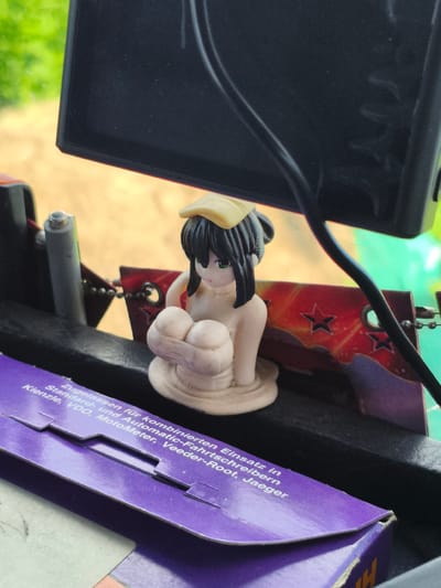 An anime figurine of a young woman with black hair and green eyes, wearing a yellow headpiece and a light-colored garment, is positioned on a black surface. The figurine is made of a creamy-white material. Behind the figurine, a red rectangular object with yellow star-like patterns and a black border is visible. To the left of the figurine, a silver cylindrical object is present. The scene appears to be indoors, with a blurry background suggesting outdoor elements like greenery and sunlight. In the foreground, a portion of a purple box with white text is visible. The text on the box includes the words "Zugelassen für," "Einsatz in," "Standard- und Automatic-Fahrtsc," "schreibern," and "Kienzle, VDO, MotoMeter, Veeder-Root, Jaeger". The image does not contain any specific information to place it in Il'mino, Russia.