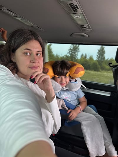 In this image, two young people are inside a vehicle. The person in the foreground is lying down, looking towards the camera with their head resting on their hand. They have dark hair and are wearing a white long-sleeved shirt. Beside them, another young person is also lying down, nestled amongst stuffed animals. This second person is wearing a blue shirt with a floral design and light gray pants. They appear to be sleeping or resting, with their face partially obscured by a giraffe-shaped stuffed animal. The scene inside the vehicle shows part of the interior, including a gray ceiling with a light fixture and a handle. Through the windows, a blurry green landscape with trees is visible, suggesting the vehicle is in motion or parked amidst nature. The lighting suggests it might be daytime, with natural light filtering in. The overall mood seems relaxed and familial, with the presence of stuffed animals adding a touch of comfort and playfulness. No visible text is present in the image.