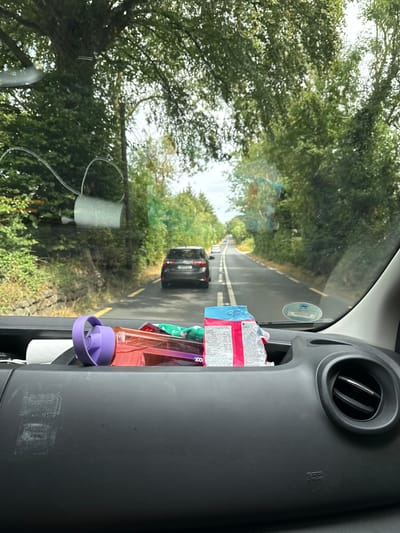 The image is taken from inside a vehicle looking through the windshield. The view is of a tree-lined road with a black SUV driving ahead. On the dashboard of the vehicle, a pink water bottle with a purple lid is visible, along with a package of cookies. A roll of toilet paper is attached to the windshield. The scene suggests the vehicle is on a journey through a rural area, possibly in the city of New Ross Municipal District, Ireland, given the lush greenery and the type of road.