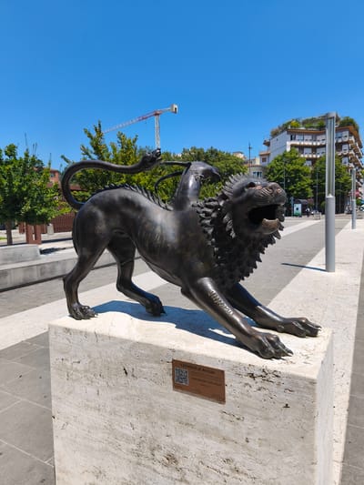 The image shows a bronze sculpture of the Chimera of Arezzo on a stone pedestal in a public square. The sculpture depicts a mythical creature with the body of a lion and a goat's head emerging from its back, with a serpent's head forming its tail. The sculpture is dark bronze and detailed, with the lion in a dynamic pose, paws outstretched, and mouth open. A QR code and text plaque are affixed to the pedestal, identifying the sculpture as "CHIMERA DI AREZZO" and crediting the "DONO ROTARY CLUB AREZZO." The background shows a clear blue sky, green trees, modern buildings with balconies and green roofs, and streetlights. In the distance, a construction crane is visible. The ground is paved with light-colored stone tiles. The scene appears to be a sunny day in a city square.