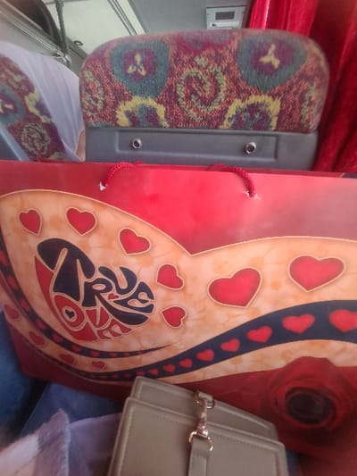 A red gift bag adorned with heart motifs and an abstract design sits beside a patterned bus seat. A small, beige purse with a gold clasp partially obscures the bag. A red curtain provides a backdrop, with the interior of the bus visible on the left.