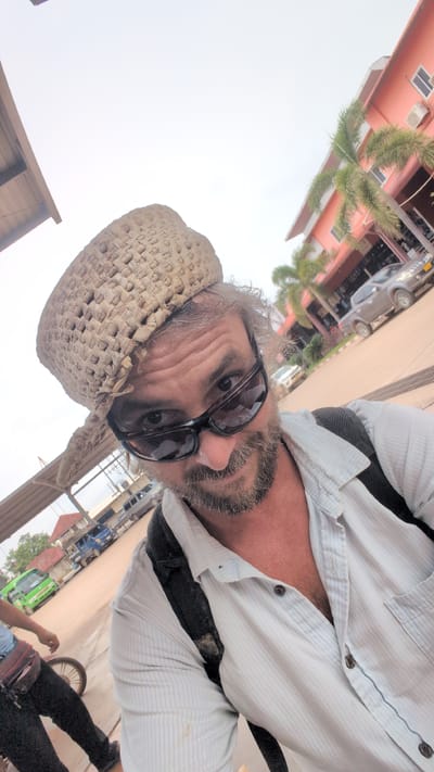 A man wearing a straw hat and sunglasses takes a selfie outdoors. He is wearing a light blue striped shirt and a dark backpack. The background shows a building with a pink facade and palm trees, as well as a street with vehicles, including a green van and several cars. The scene appears to be in Phakhao, Laos.