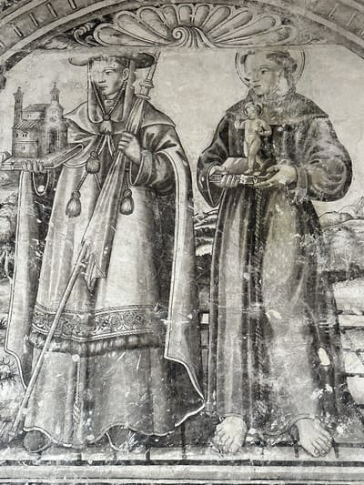 The image is a fresco depicting two figures in religious attire. The figure on the left wears a wide-brimmed hat and a hooded robe with decorative tassels and a patterned belt. This figure holds a staff in their right hand and what appears to be a miniature church or building in their left hand. To the left of this figure is a depiction of a two-towered church with a cross on top. The figure on the right wears a hooded robe and a halo around their head. This figure holds a book in their left hand and a small naked child with a cross in their right hand. The background of the fresco includes a landscape with trees and hills. The fresco is located in the *Ex-convento de San Miguel Arcángel* in Huejotzingo, Mexico.