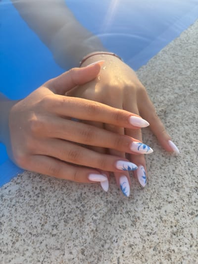 In Delchevo, North Macedonia, a close-up shot focuses on a pair of hands with almond-shaped, light pink nails, several of which are adorned with delicate blue floral decals. The hands are submerged in bright blue water, with water droplets glistening on the skin, suggesting a swimming pool or similar aquatic environment. The hands rest on a textured, light-colored surface, possibly the edge of the pool. The overall lighting indicates a sunny day, with sunlight casting reflections on the water and highlighting the texture of the hands and the pool edge.
