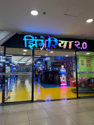 The image shows the entrance to "Zingeria 2.0", an arcade or entertainment center. The sign for Zingeria 2.0, with multicolored lettering, is prominent over the entrance. Inside, there are arcade games, including a Batman-themed game, a car racing game, and other machines with flashing lights. The floor is a combination of yellow and blue. The reflection shows the tile flooring. One or two people are visible inside the arcade. The location is likely in Kalyan-Dombivli, India.