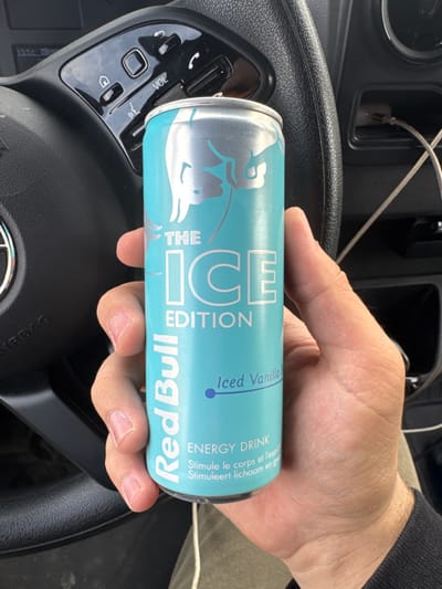 A human hand holds a light blue Red Bull "THE ICE EDITION" "Iced Vanilla" energy drink can, positioned centrally in the frame. The can features silver Red Bull branding and text in English, French, and Dutch. The hand is that of an adult, with visible fingernails. The background reveals the interior of a vehicle. A black multi-function steering wheel, likely belonging to a Toyota given the partial logo visible on the airbag cover, occupies the left and upper-middle portions of the image. Steering wheel controls, including volume (VOL) and call buttons, are visible. In the upper left corner of the dashboard, a digital display shows "13:51." To the right of the steering wheel, a dark plastic dashboard section shows a USB port with charging cables connected. The specific location within the city of Roeselare, Belgium, is not discernible from the visual information provided.