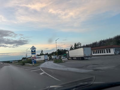 The image depicts a roadside scene near Kalugerovo, Bulgaria, under a twilight sky with scattered clouds. In the foreground, a paved road curves to the left, suggesting a highway or main thoroughfare. To the right of the road, there's a gas station with a prominent sign displaying fuel prices and operating hours ("24/7"). Several large white semi-trailer trucks are parked or driving in the vicinity of the gas station and a low-rise building with a red-tiled roof. The landscape behind the gas station appears to be a mix of trees, some of which show signs of fire damage, and the sky is a gradient of pale blue, pink, and white. The overall impression is one of a commercial rest stop on a highway at dusk.