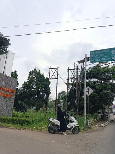 A woman in a black outfit and helmet sits on a white scooter next to a road. To the left, "Universitas" is partially visible on a stone wall. Behind her are tall trees, utility poles with wires, and a green sign indicating "POLRES BANDUNG PUSDIK POLRI" and "POLSEK KATAPANG" with arrows pointing right. A road sign with a "T" intersection symbol is also visible. The sky is overcast.