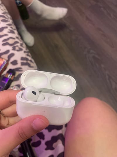 A person's hand is holding an open white wireless earbud charging case. One earbud is inside the case. The background shows a person's legs wearing white socks and a patterned surface. The location is not determinable from the image and cannot be specified as Romanovka, Russia.