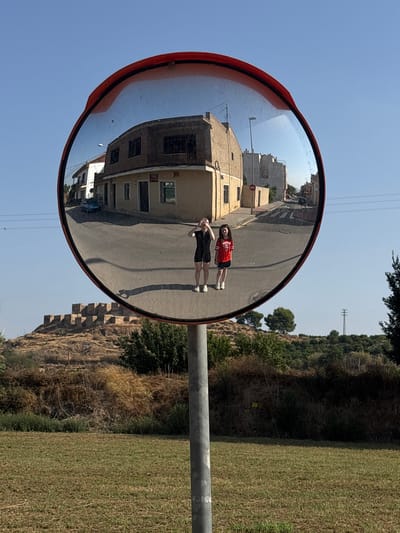 The image is a wide-angle reflection in a convex mirror, positioned on a metal pole in what appears to be a rural or semi-rural area. The mirror is framed with red. Reflected in the mirror is a street scene in the city of Monteagudo, Spain. Two young girls are visible in the reflection, standing on the asphalt road. One girl, on the left, is wearing a black outfit and holding a phone up, taking a picture. The other girl, on the right, is wearing a red t-shirt with white lettering and white shorts. They are positioned in front of a light-colored building with multiple windows and a dark-tiled roof. Further down the street, more buildings, a blue car, and streetlights are visible. Beyond the immediate street scene in the reflection, a barren, hilly landscape with some vegetation and what appear to be ancient stone fortifications or ruins are visible. The sky in the reflection is a clear, bright blue. In the foreground of the actual image, outside the mirror's reflection, is a grassy field.