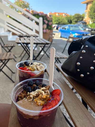 Two acai bowls in clear plastic cups are prominently displayed on a wooden slatted outdoor table in Ribamontán al Mar, Spain. The front bowl contains a dark purple base, a layer of light brown spread, blueberries, sliced strawberries, granola, slivered almonds, and a yellow, seedy fruit pulp, with a small blue flower garnish. A beige compostable spoon is upright in this bowl. The second bowl, positioned behind the first, also features a purple base, a light brown spread, blueberries, sliced strawberries, granola, slivered almonds, and sliced bananas, with an identical compostable spoon. A black leather handbag adorned with numerous silver studs is partially visible on the table to the right. The background consists of an outdoor dining area with additional wooden tables and chairs, a white wooden railing, and pink flowering plants. Beyond this area, a blurred street scene shows a bright blue hatchback car and other vehicles. Residential buildings with terracotta roofs and green foliage are visible in the distant background under a clear blue sky. No individuals are directly depicted.