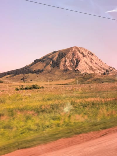 The image captures a landscape scene, likely from a moving vehicle, given the motion blur in the foreground. A prominent, light-colored, rounded mountain dominates the mid-ground. The mountain's slopes are partially covered with sparse dark vegetation, and its upper portion appears eroded with visible striations. In front of the mountain, a rolling landscape of dry grass and scattered bushes stretches across the frame. A few thin, vertical poles, possibly fence posts, are visible in the grassy area. The sky above is a pale gradient of blue and pink, with a thin, dark horizontal line, possibly a power line or wire, crossing the upper part of the image. The foreground exhibits significant motion blur, showing streaks of green and reddish-brown, suggesting a road or path moving past. The scene appears to be an outdoor natural environment.
