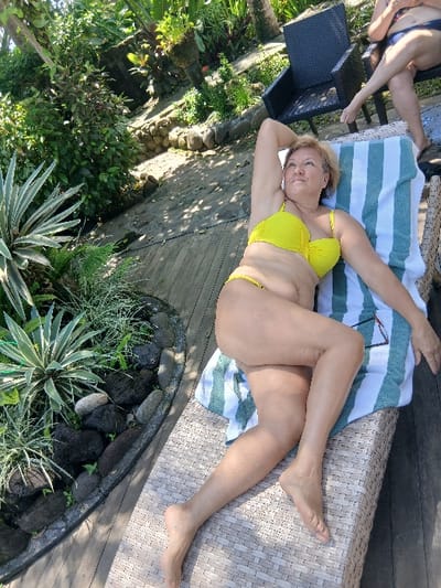 The image displays an outdoor scene in Tibubiyu, Indonesia, featuring two individuals on a wooden deck adjacent to a tropical garden. In the foreground, a fair-skinned, middle-aged woman with light hair is reclined on a light-brown woven lounge chair, wearing a bright yellow two-piece bikini. Her right arm is bent with her hand near her head, and she is looking upwards and to her left. A white towel with green stripes is draped over the lounge chair, with a pair of sunglasses (black frame, red temples) resting on it next to her left arm. In the upper right background, a second, partially visible individual is seated on a dark woven chair, facing away from the camera, wearing a dark bikini and with legs crossed. The environment consists of dense green foliage, including succulents and ferns, bordered by dark rocks. Potted plants are also visible in the background. The scene is illuminated by natural light, casting shadows on the wooden deck.
