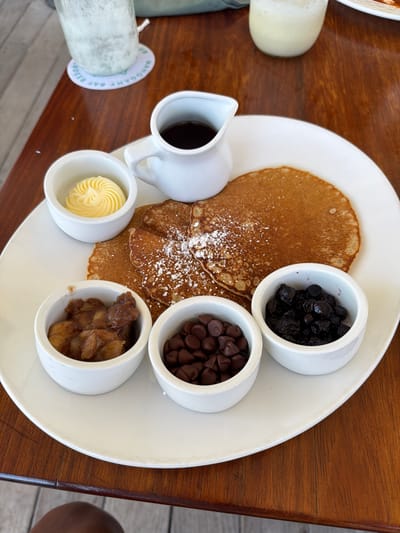 A high-angle shot captures a breakfast setting on a wooden table at the Mahogany Bay Resort in San Pedro Town, Belize. The central subject is a large white oval plate holding several golden-brown pancakes, lightly dusted with powdered sugar. Surrounding the pancakes are five small white ramekins containing various toppings: a swirl of light yellow butter in the upper left, a small white pitcher filled with dark brown syrup to its right, cooked apple pieces or compote in the lower left, milk chocolate chips in the lower middle, and dried blueberries in the lower right. In the upper left background, a tall glass containing a light, frothy beverage rests on a white circular coaster clearly marked "MAHOGANY BAY RESORT" and "SAN PEDRO, BELIZE." The table surface is a reddish-brown wood with distinct grain, and a small portion of a person's dark-skinned thigh is visible in the bottom left corner of the frame.