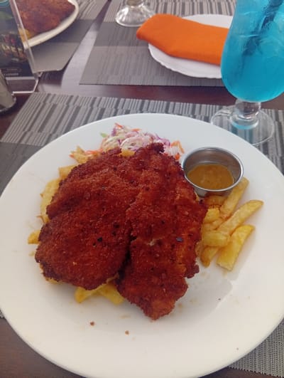 A detailed view of a dining table in Murang'a, Kenya, presents a served meal within a restaurant setting. In the foreground, a white oval plate features three pieces of deep reddish-brown breaded cutlets, surrounded by golden-brown French fries and a small portion of coleslaw comprised of shredded red and white cabbage with carrots. A small silver ramekin on the plate contains a light brown, viscous sauce. To the right, a stemmed glass holds a vibrant blue beverage with a visible straw. These items are positioned on a grey-and-white striped placemat, which rests on a dark brown wooden table. In the background, another identical placemat is set with a white plate, a folded orange cloth napkin, and an empty stemmed glass. A partial view of a clear acrylic menu stand and an additional white plate holding a similar dark brown, breaded item is visible on the far left. No individuals are present in the scene.