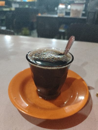 An overhead-angle image depicts a clear glass filled with dark brown liquid, consistent with coffee, approximately 90% full, topped with a layer of small bubbles. A reddish-brown plastic spoon is inserted vertically into the liquid. The glass rests centrally on a glossy, circular orange saucer. Both objects are positioned on a flat, light-colored surface, likely a tabletop, casting soft shadows to the right. The background is blurred and out of focus, revealing indistinct shelves or a counter with various containers in an indoor setting, likely a commercial establishment such as a cafe or warung in Paloh Teungoh, Indonesia. No individuals are visible.