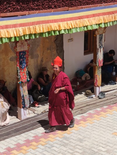 In the city of Pibiting, India, a Buddhist monk in a red robe and hat walks on a cobblestone path. The monk is wearing glasses and appears to be smiling. Behind the monk, a building with a yellow facade and a colorful canopy is visible. Several other people are seated on the ground near the building, some of whom are also wearing traditional attire. The scene is lit by bright sunlight, casting shadows on the ground.