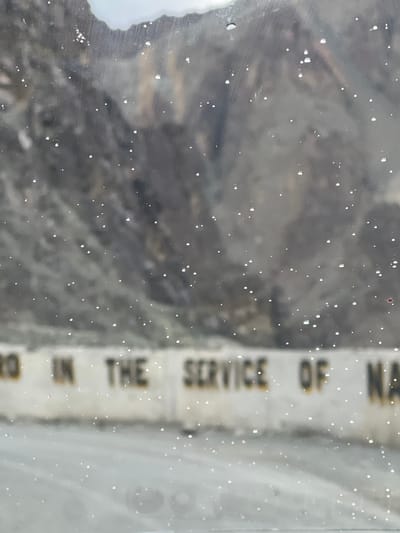 The image shows a blurred view of a mountainous landscape through a surface with numerous water droplets and small white specks. In the foreground, a low, white wall with yellow lettering is visible. The visible text on the wall reads, "IN THE SERVICE OF NA". The wall appears to be a guardrail or retaining wall, likely along a road. The background consists of blurred, rocky mountains with patches of light and shadow. The overall impression is of a scene viewed from inside a vehicle on a wet or misty day, possibly in a mountainous region. The text suggests a patriotic or service-oriented message, common on infrastructure in certain locations. The location in Khaltse, India, would imply a mountainous terrain, possibly along a road like the Leh-Srinagar highway.