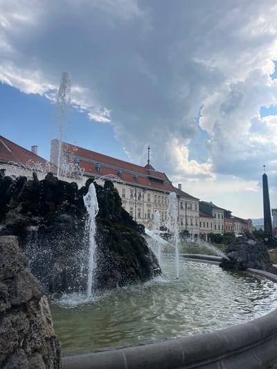 The image depicts a close-up view of a fountain in front of a row of historic buildings in Banská Bystrica, Slovakia. The fountain features a rocky, moss-covered structure with water spraying upwards and outwards, creating a misty effect. In the background, several buildings with red tiled roofs and ornate facades line a street. To the far right, a tall, dark obelisk with a star on top stands against the cloudy sky. The sky itself is a mix of blue patches and large, dramatic grey clouds. The foreground is dominated by the fountain and its basin, with the buildings and obelisk forming the backdrop. There are no people visible in the image.