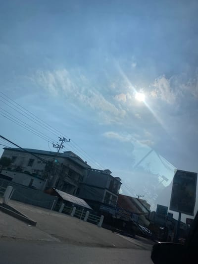 The image captures a street scene in Kertak Hanyar I, Indonesia, under a bright, partly cloudy sky. The sun is prominently visible, casting a strong glare and lens flare. In the foreground, a paved road curves to the left, with a concrete barrier along its edge. Beyond the road, a row of buildings lines the street. One building, appearing to be a commercial establishment, has signs with text that includes "SINAR MOTOR SHOW ROOM" and "MURAH VARIASI MOBIL". Another building to the left is a multi-story structure with visible windows and a faded facade. Overhead, numerous power lines crisscross the frame, supported by utility poles. The sky is a clear blue with scattered white clouds. A reflection of a pyramidal structure is visible on the surface that captured this image, possibly a window or windshield. A large billboard stands on the right side of the street, displaying a picture of a person. The overall impression is of a typical urban street in Indonesia on a sunny day.