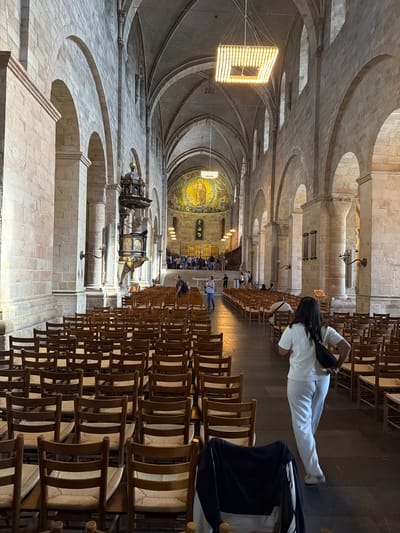 The image shows the interior of Lund Cathedral in Lund, Sweden. The camera is positioned looking down the nave of the cathedral towards the apse. Rows of empty wooden chairs with woven seats fill the floor. The cathedral's architecture is characterized by stone walls, arched ceilings, and large pillars. A woman in white pants and a white t-shirt with a black crossbody bag is walking away from the camera down the center aisle. In the background, there are a few people gathered near the altar area, which is adorned with a large mosaic. A pulpit is visible on the left side of the nave. The lighting appears to be a mix of natural light coming from high windows and artificial light from a large, modern chandelier and wall sconces.