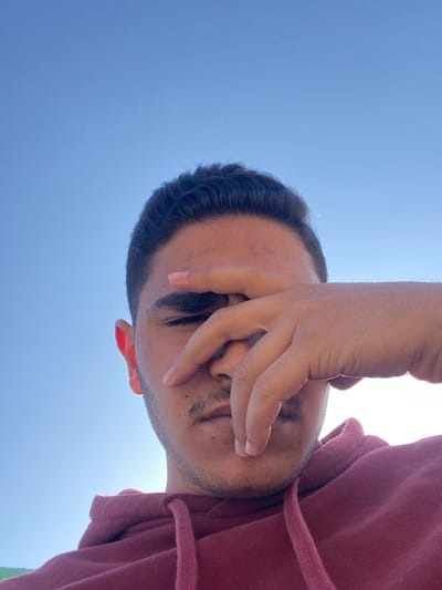A man with dark hair is captured from a low angle, with the blue sky as the backdrop. He partially covers his face with his hand, shielding his eyes from the bright sunlight. He wears a maroon hoodie. The composition focuses on the contrast between the man's features and the stark blue background.