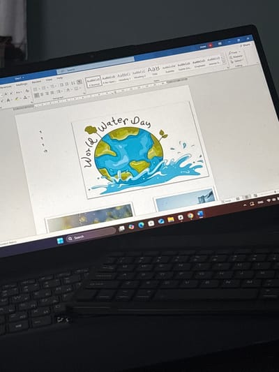 The scene depicts a dark-colored laptop displaying a Microsoft Word document, located in the city of Nadarevo, Bulgaria. The laptop screen shows a central illustration of the Earth surrounded by splashing water, accompanied by the text "World Water Day". Below this, smaller image thumbnails are visible within the document or application interface. The Microsoft Word ribbon menu is active at the top of the screen, displaying standard options, with the document titled "Doc1". The Windows 11 taskbar is present at the bottom of the screen, showing various pinned application icons including web browsers and utility tools, along with system tray icons indicating an English language setting. The top portion of the laptop's keyboard is visible below the screen, featuring a QWERTY layout with visible Cyrillic characters, suggesting a Bulgarian or dual-language keyboard. The background environment is indistinct and dimly lit. No individuals are visible.