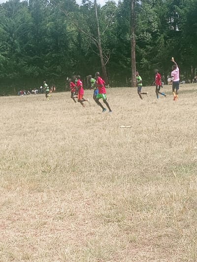 This image depicts an outdoor football match in Lwandeti ward, Kenya. The scene features a large, open field covered in dry, brownish-green grass, backed by a dense line of tall green trees. Ten individuals, primarily young males, are visible across the field. Seven players are in active motion, engaged in the football game, wearing either red or green sports shirts with dark shorts. On the far right, one player in a light pink shirt and dark shorts holds a light-colored football aloft with his right hand. In the distant left background, a small group of people are seated near the treeline, observing the match. The sky is overcast.