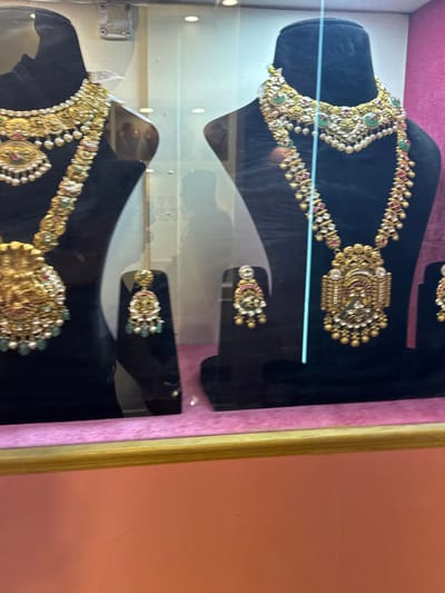 The image displays a retail jewelry showcase in Jaipur Municipal Corporation, India. Two black velvet jewelry busts are featured behind a clear glass panel. The left bust exhibits a multi-layered gold necklace with an ornate central pendant depicting a deity, adorned with pearls and green gemstones, accompanied by a matching pair of earrings on a smaller stand. The right bust showcases a multi-layered gold necklace with a large, rectangular central pendant, embellished with red and green gemstones, white stones, and pearls, also with a corresponding pair of earrings on an adjacent stand. The base of the display case is lined with pink velvet fabric, and a golden wooden frame borders the bottom edge. Reflections of individuals and interior shop elements are faintly visible on the glass surface. The background wall behind the display is a light pink or coral hue. No explicit human actions or interactions are depicted.