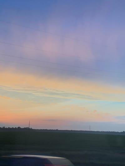 The image captures a serene, dusky sky over a rural landscape. The sky transitions from a deep blue at the top to softer hues of pink, orange, and pale blue near the horizon, suggesting either sunrise or sunset. Wispy clouds are visible, tinged with the colors of the sky. Several faint, parallel lines, likely power lines, stretch horizontally across the upper portion of the sky.

Below the sky, a dark, silhouetted horizon line reveals a dense line of trees and vegetation. In front of this, a large, dark green field occupies the middle ground, possibly a cultivated field or pasture. The foreground shows the edge of a dark surface, possibly a road or car, with a faint red reflection on it. Two solitary utility poles stand in the field, one more prominent than the other, breaking the monotony of the landscape. The overall impression is one of quietude and natural beauty.

There is no information available to determine the specific location within the city of Viktoriia, Ukraine.