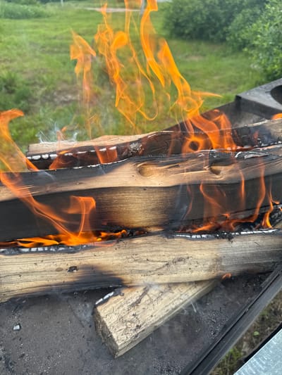 The image displays a close-up, outdoor shot of a campfire. Several pieces of wood are stacked in a fire pit, with bright orange flames consuming them. The wood appears to be dry and aged, with some sections already blackened by the fire. Wisps of white smoke are visible curling up from the burning logs. The background is blurred but suggests a grassy area with green foliage, hinting at a natural or park-like setting. The fire pit itself is made of dark metal. The overall scene evokes a sense of warmth and the primal act of burning wood. The location is identified as Луптюг, Russia.