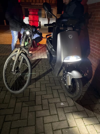 At night in Holtzheim, France, two individuals are present on a paved surface consisting of dark gray herringbone-patterned pavers. On the left, a person wearing a dark jacket and blue jeans stands near a dark-colored bicycle, which has its front light illuminated. A bag is attached to the bicycle's handlebars. On the right, a second individual, dressed in a dark jacket, sits on a silver-gray Yadea electric scooter, its headlight actively illuminated. The scooter is positioned close to a red brick wall. In the background, behind both vehicles, a stack of large, rectangular objects is heavily illuminated by a red light source, casting a strong red glow onto the surrounding area. The scene is lit by the vehicles' headlights and the ambient red light, creating shadows on the ground and the adjacent wall.