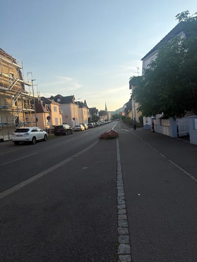 This is a medium-angle, full-shot image of a street in Singen (Hohentwiel), Germany, taken from a low perspective. The scene captures a residential street with houses on both sides and cars parked along the curb. In the foreground, a dark asphalt road is visible with a border of light-colored cobblestones running along its edge. A sidewalk made of the same cobblestone material is to the right.

On the left side of the street, several parked cars are visible, including a white Lexus SUV in the foreground. Behind the cars, there are buildings with red-tiled roofs and light-colored facades. One building on the far left is partially covered in scaffolding, suggesting ongoing construction or renovation.

On the right side, more houses with similar architectural styles are present. A large tree with lush green foliage extends into the frame, casting a shadow on the sidewalk. A white picket fence lines a portion of the property. A single individual, a man dressed in a gray jacket and dark pants, is walking away from the camera on the sidewalk. Further down the street, a red fire hydrant is visible near the fence.

In the distance, the street curves gently, and a church spire can be seen rising above the rooftops. The sky is clear and blue with a hint of haze near the horizon, indicating it is likely late afternoon or early evening. The overall impression is a quiet, suburban street scene.