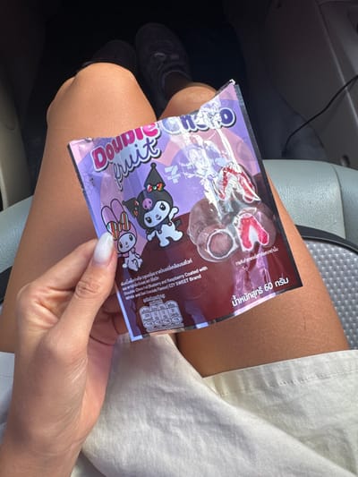 A person's hand with manicured nails holds a purple and maroon snack package in their lap. The package has text in Thai and English, featuring cartoon characters that appear to be from the Sanrio universe, specifically My Melody and Kuromi. The snacks inside are visible through the clear plastic packaging, appearing to be chocolate-covered fruit pieces. The background shows the interior of a vehicle, with a gray seat visible. The person's legs are also in the frame, suggesting they are seated in the car. The scene implies a casual moment, possibly during travel, in Ban Huai Phlap, Thailand.