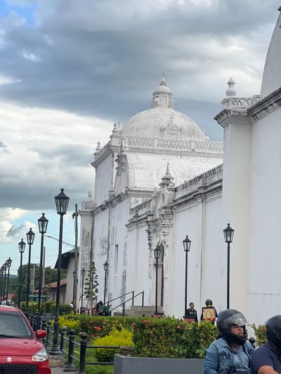 The image depicts a street view in the city of León, Nicaragua, dominated by a large, white colonial-era building that appears to be a church or cathedral. The building features a prominent dome and ornate architectural details, with some signs of aging on its facade.

In the foreground, a red Suzuki car is partially visible on the left. A row of black lampposts lines the street. Between the lampposts and the building is a well-maintained garden with lush green bushes and small red flowers.

Several people are present in the scene. On the steps of the building, a person wearing a green shirt is seated. In the mid-ground, two individuals are standing, one of whom is wearing a black jacket with a yellow box on their back, emblazoned with a golden arch logo, suggesting a food delivery service. In the immediate foreground, two individuals wearing helmets are seated on a motorcycle, with one facing the camera and looking to the side.

The sky is cloudy with patches of blue sky visible. The overall atmosphere suggests an overcast day in an urban environment. The architecture and the presence of people in casual attire point to a typical street scene in a historic Central American city.