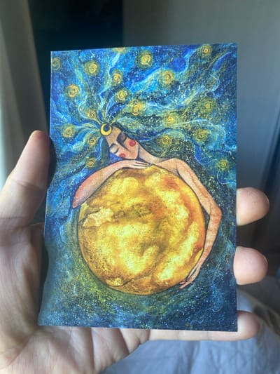A person's hand holds a postcard featuring an artwork. The artwork depicts a stylized figure with long, flowing blue and yellow hair adorned with small yellow circular motifs. The figure has closed eyes and is cradling a large, round, yellow object, possibly the sun or moon. The background of the artwork is a deep blue with scattered yellow specks and swirling patterns, suggesting a starry night or cosmic scene. The postcard itself appears to be slightly creased. The location is not visible in the image.