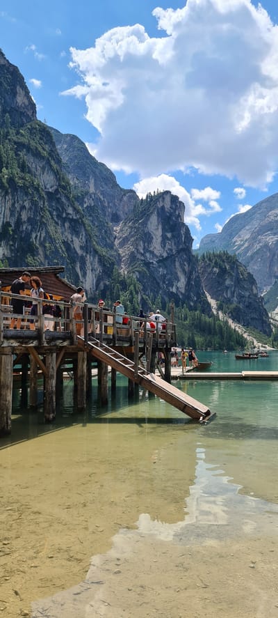The image depicts a scenic view of a wooden dock extending into a clear, greenish-blue lake, with imposing, rugged mountains and lush green trees in the background. The sky is a vibrant blue, adorned with fluffy white clouds, suggesting a pleasant, sunny day, likely late morning or early afternoon.

Several people are visible on the dock, some appearing to be tourists enjoying the view or preparing for an activity. They are dressed in casual, summer attire, with a mix of hats and t-shirts. A few individuals are seated or standing, some leaning over the railings to look at the water or the distant scenery. The dock itself is constructed of weathered wood, with sturdy posts supporting its structure and a ramp leading down to the water. In the distance, small boats, possibly rowboats or canoes, can be seen on the lake, with people in them.

The water is remarkably clear, allowing the sandy, pebble-strewn lakebed to be visible in the shallows near the dock. The reflection of the sky and the dock is partially visible on the water's surface. The mountains are dramatic and imposing, with visible layers of rock and patches of green vegetation and trees clinging to their slopes. The overall impression is one of a serene and beautiful natural landscape, likely a popular tourist destination.

There are a few small signs on the dock, but the text is not legible. The mood conveyed is one of leisure and appreciation for the natural beauty of the location. The orientation of the image is vertical, as if captured while standing and looking up towards the mountains and sky.