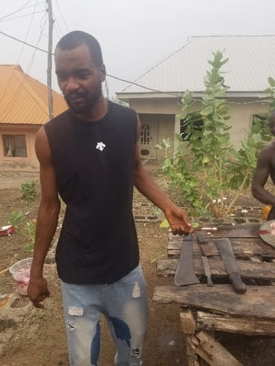 The image shows a man standing in front of a wooden table with three large knives laid out on it. He is wearing a black tank top and blue jeans with rips in the knees. He is looking at the camera with a serious expression. The table is situated in a yard that is partially paved and partially covered in dirt. Behind the man are two partially constructed buildings with corrugated metal roofs.  One is tan and the other is grey. There is a small tree in the background and another with green leaves to the right of the man. The man's left hand is resting on the table, the right is held up as if pointing at the knives. The man's upper body is turned slightly to the left. Behind the man in the distance, a partial view of another individual is visible, standing behind a portion of the grey building and is facing away from the camera. The location is in Pati Ridge, Nigeria, at latitude: 7.81, longitude: 6.704.  The scene is somewhat casual, but there is an implied sense of tension.