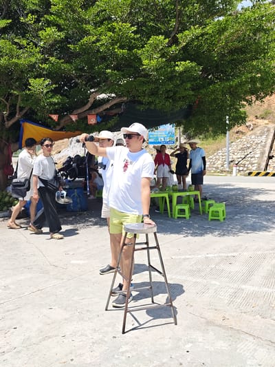 In the city of Vinh Hai, Vietnam, a group of individuals are gathered under the shade of a large tree. The scene appears to be a casual outdoor gathering, possibly a resting spot or a small market.
In the foreground, a man wearing a white t-shirt, white baseball cap, and yellow shorts is standing on a metal stool, holding a camera and pointing it to his right, as if taking a photo or filming. He is looking in the same direction. Beside him, to his left, another man in a white t-shirt and black pants is walking, holding a silver helmet.
Further back, a group of people are standing near green tables and stools, with several coconuts visible on the tables. One woman is wearing a red shirt and white shorts, another is in a dark blue t-shirt and black skirt, and a man is in a light blue t-shirt and dark shorts. They are all wearing straw hats. Another man in a white t-shirt and white shorts is walking towards the left, carrying a black bag.
Behind the people and tables, motorcycles are parked, and blue bags are piled on the ground. A yellow and orange tarp is stretched out under the tree. The ground is paved with concrete, and a slight incline is visible in the background, with a stone wall and a concrete structure. Sunlight filters through the leaves of the tree, creating dappled shadows on the ground. The overall impression is a relaxed, sun-drenched day in a semi-tropical environment.