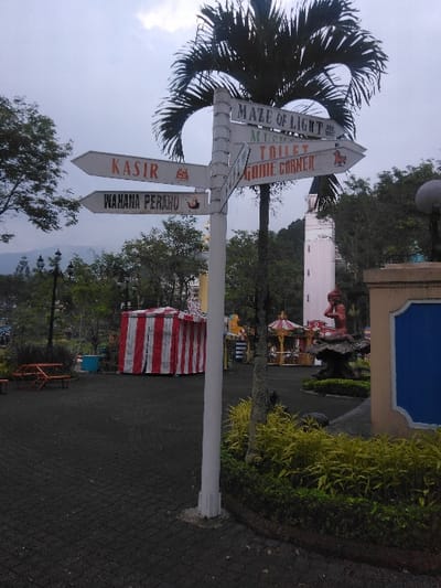 The image depicts an outdoor amusement or park area. A prominent white, multi-armed directional signpost stands centrally, fixed to a pole beneath a large palm tree. The signpost features six directional arrows. Clockwise from the upper left, the visible labels read: "KASIR" (Cashier) with a book icon; "WAHANA PERAHU" (Boat Ride) with a pipe icon; "MAZE OF LIGHT"; "MICROS..." (partially obscured); "TOILET" with a figure icon; and "GAME CORNER" with a horse icon. The language on "KASIR" and "WAHANA PERAHU" is Indonesian, indicating the location is in an Indonesian-speaking region, not Ciudad de San José, Costa Rica.
The ground in the foreground consists of dark gray paved blocks. To the immediate right of the signpost, a manicured garden bed with green and yellow foliage is visible. In the midground to the left, a large red and white striped tent structure is present, along with wooden benches. Further back on the right, an ornamental statue of a reddish-brown figure (possibly a mermaid) on a dark base is positioned near a blue and beige structure. Behind the garden, a carousel ride with a red and white striped canopy is partially visible. In the far background, a tall, light-colored tower with a pointed roof rises above trees, and distant mountains are faintly discernible under an overcast sky. No individuals are visible. The sky is uniformly light grey, suggesting an overcast day.
