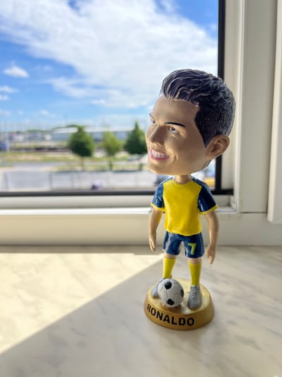 A Cristiano Ronaldo bobblehead is displayed on a marble windowsill, facing left. The figurine wears a yellow jersey with blue trim and dark blue shorts with the number 7. It stands on a gold base inscribed with "RONALDO" and has a soccer ball at its feet. The background, seen through a window, shows a blurred urban landscape with trees, buildings, and a cloudy blue sky. Sunlight casts a shadow across the windowsill and the figurine, creating a diagonal line of light and shade. The scene is set in Kobylnica, Poland.